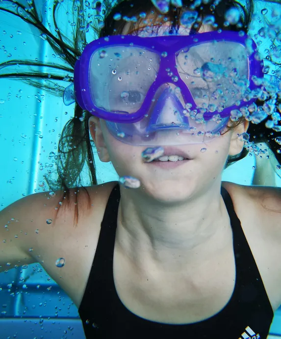 Kind taucht mit lila Taucherbrille im türkisblauen Hallenbad-Pool unter Wasser